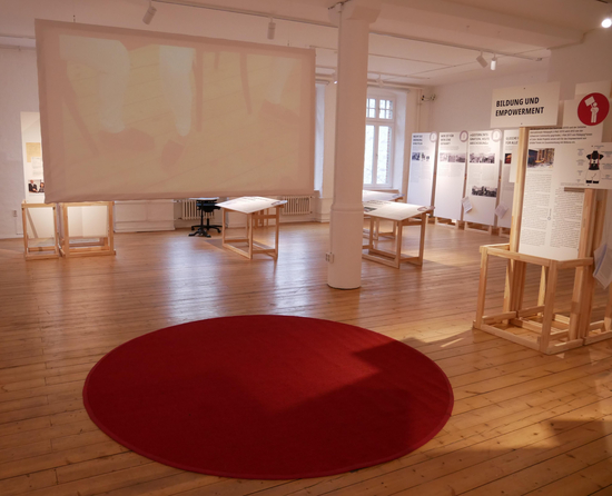 Ausstellungsraum mit Holzfußboden und rundem rotem Teppich. Im Hintergrund sind Ausstellungstafeln zu erkennen.